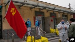 2022年3月18日，北京居民社区工作人员身穿防护服站在一堆标有“有害物品”的黄色塑料袋旁交谈。后面墙上挂着一面中共党旗。