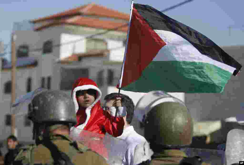 Seorang bocah Palestina mengenakan pakaian Santa Claus memegang bendera Palestina berdiri di depan polisi perbatasan Israel dalam demonstrasi menentang dipasangnya penghalang pemisah di desa Al-Masara, Tepi Barat, dekat Betlehem.