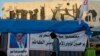 An anti-government protester walks by a defaced picture of Prime Minister-designate Adnan al-Zurfi in Tahrir Square, Baghdad, Iraq, March 19, 2020.