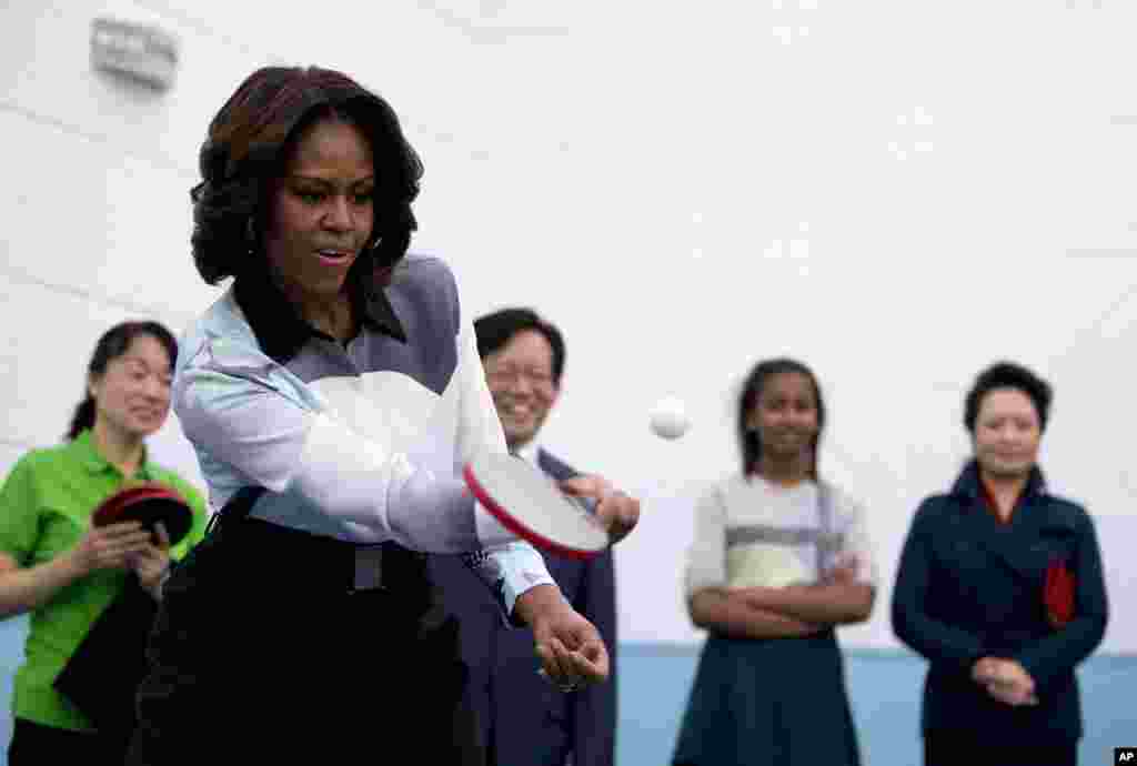Ibu Negara Amerika Michelle Obama bermain tenis meja sementara Peng Liyuan, istri Presiden Cina Xi Jinping (berdiri paling kanan), dan yang lainnya memperhatikannya dalam kunjungan mereka ke sebuah sekolah di Beijing yang mempersiapkan para siswa menuju perguruan tinggi di luar negeri. 