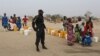 FILE - A Cameroonian police officer stands next to people waiting for water at the Minawao refugee camp for Nigerians who fled Boko Haram attacks in Minawao, Cameroon, March 15, 2016.