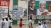 Street-vendors and others walk past a billboard showing Nigeria's President Muhammadu Buhari and other party officials, in Kano, northern Nigeria, Feb. 26, 2019. Buhari's election lead grew on Tuesday.