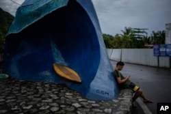 A resident sits by a sculpture of the Teahupo'o wave at the end of the road, Tahiti, French Polynesia, Saturday, Jan. 13, 2024. (AP Photo/Daniel Cole)