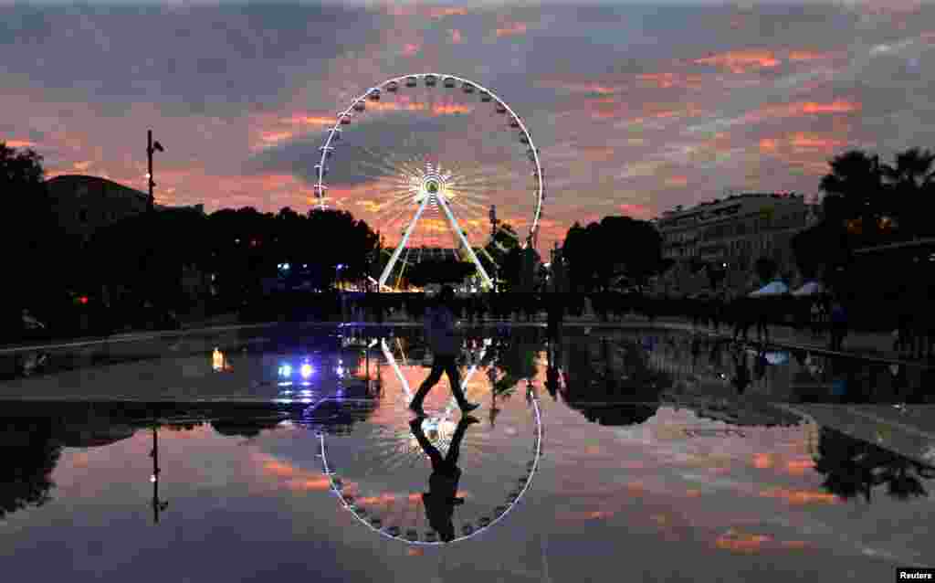 បុរស​ម្នាក់​ដើរ​​​ដោយ​មាន​រូប​ឆ្លុះ​ពី​អាង​បាញ់​ទឹក ស្របពេល​ខ្លួន​ដើរ​កាត់​ទោង​វិល Ferris Wheel អំឡុងពេល​ពិព័រណ៍​ភ្លើង​ពណ៌​ប្រចាំ​រដូវ​ឈប់សម្រាក​ថ្ងៃ​បុណ្យ​ណូអែល ក្នុង​ក្រុង Nice ប្រទេស​បារាំង។&nbsp;