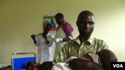 Savio Niwagaba holds his newborn baby as his wife Chrisente, behind him, has a contraceptive implant inserted in her arm, Kanungu, Uganda, June 19, 2012. (VOA / Hilary Heuler)
