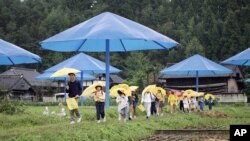 ບັນດາເດັກນ້ອຍນັກຮຽນຂອງໂຮງຮຽນລັດ ຖື​ຄັນ​ຮົ່ມ​ສີ​ເຫຼືອງ​ຂອງ​ຕົນ​ເອງ​ຍ່າງຜ່ານ​ທົ່ງ​ນາບ່ອນ​ທີ່​ມີຄັນ​ຮົ່ມ​ສີ​ຟ້າ​ຂະ​ຫນາດ​ໃຫຍ່​ຂອງ​ໂຄງ​ການ​ໂດຍ ຄຣິສໂຕ ໃນ ຮ່ອມພູ ແມ່ນໍ້າ ຊາໂຕະ ຂອງ​ຍີ່​ປຸ່ນ​, ວັນ​ທີ 9 ຕຸ​ລາ 1991. (ຮູບພາບຈາກ ອົງການຂ່າວ AP ພາບ /ໂດຍ ມິຊູຮິໂກະ ຊາໂຕະ (Mitsuhiko Sato))