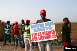 An African migrant laborer holds a banner reading "against all ghetto, for the dignity of work" during a march to protest against his work conditions in Italy, following the death of 16 colleagues in two separate road accidents, near Foggia, Italy, Aug. 8, 2018.