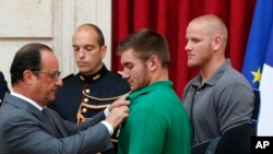 法国总统奥朗德把法国荣誉军团勋章别在美国国民警卫队成员阿列克·斯卡拉托斯身上，美国空军士兵斯宾塞·斯通在旁观看。（2015年8月24日）