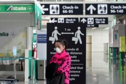 A woman wearing a protective mask stands in front of a notice recommending to keep the social distance of 1 meter between people, at the Leonardo da Vinci international airport, in Rome, March 12, 2020.