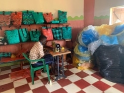 A tailor sits next to a display shelf in the Reform Africa workshop, in Mpigi district, Uganda. (H.Athumani/VOA)