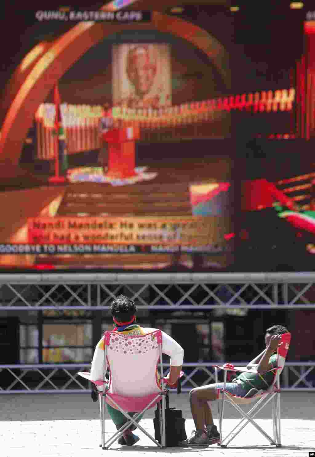 Dua orang mengikuti jalannya upacara pemakaman Nelson Mandela di layar besar di lapangan Nelson Mandela di Sandton, Johannesburg.