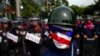 FILE - An anti-government protester wearing a mask painted in the colors of the Thai national flag looks on as riot police officers stand guard outside the parliament in Bangkok, Aug. 7, 2013. 