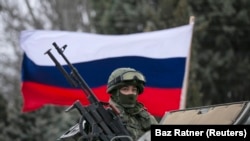 Seorang pria pro-Rusia (tidak terlihat) memegang bendera Rusia di belakang seorang prajurit bersenjata di atas kendaraan tentara Rusia di luar pos penjaga perbatasan Ukraina di kota Balaclava, Krimea, 1 Maret 2014. (Foto: REUTERS/Baz Ratner)
