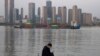 In this Monday, April 13, 2020, photo, a man wearing a mask against the new coronavirus checks his phone as a container ship cruises along the Yangtze River in Wuhan in Central China's Hubei province. China's exports fell further in March compared…