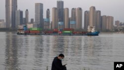 In this Monday, April 13, 2020, photo, a man wearing a mask against the new coronavirus checks his phone as a container ship cruises along the Yangtze River in Wuhan in Central China's Hubei province. China's exports fell further in March compared…