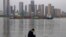 In this Monday, April 13, 2020, photo, a man wearing a mask against the new coronavirus checks his phone as a container ship cruises along the Yangtze River in Wuhan in Central China's Hubei province. China's exports fell further in March compared…