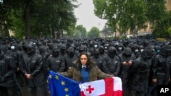 Një grua mban flamujt e Gjeorgjisë dhe BE-së para kordonit policor që ka bllokuar hyrjen e Parlamentit për të penguar protestuesit t'i afrohen godinës, 14 maj 2024, Tbilisi, Gjeorgji