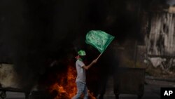 Palestinski demonstrant s Hamasovom trakom na glavi nosi zastavu Hamasa iza zapaljenih guma tokom sukoba nakon demonstracija solidarnosti s pojasom Gaze, u gradu Ramallah na Zapadnoj obali, petak, 27. oktobra 2023. (AP Photo/Nasser Nasser)